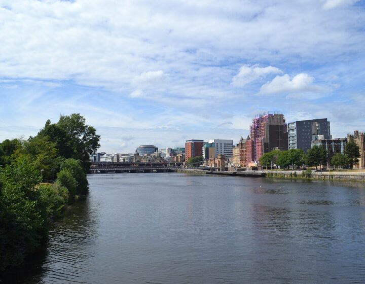 The River Clyde in central Glasgow on a bright July day, with trees in full leaf on one bank, city buildings along the other and railway bridges downstream under a blue sky