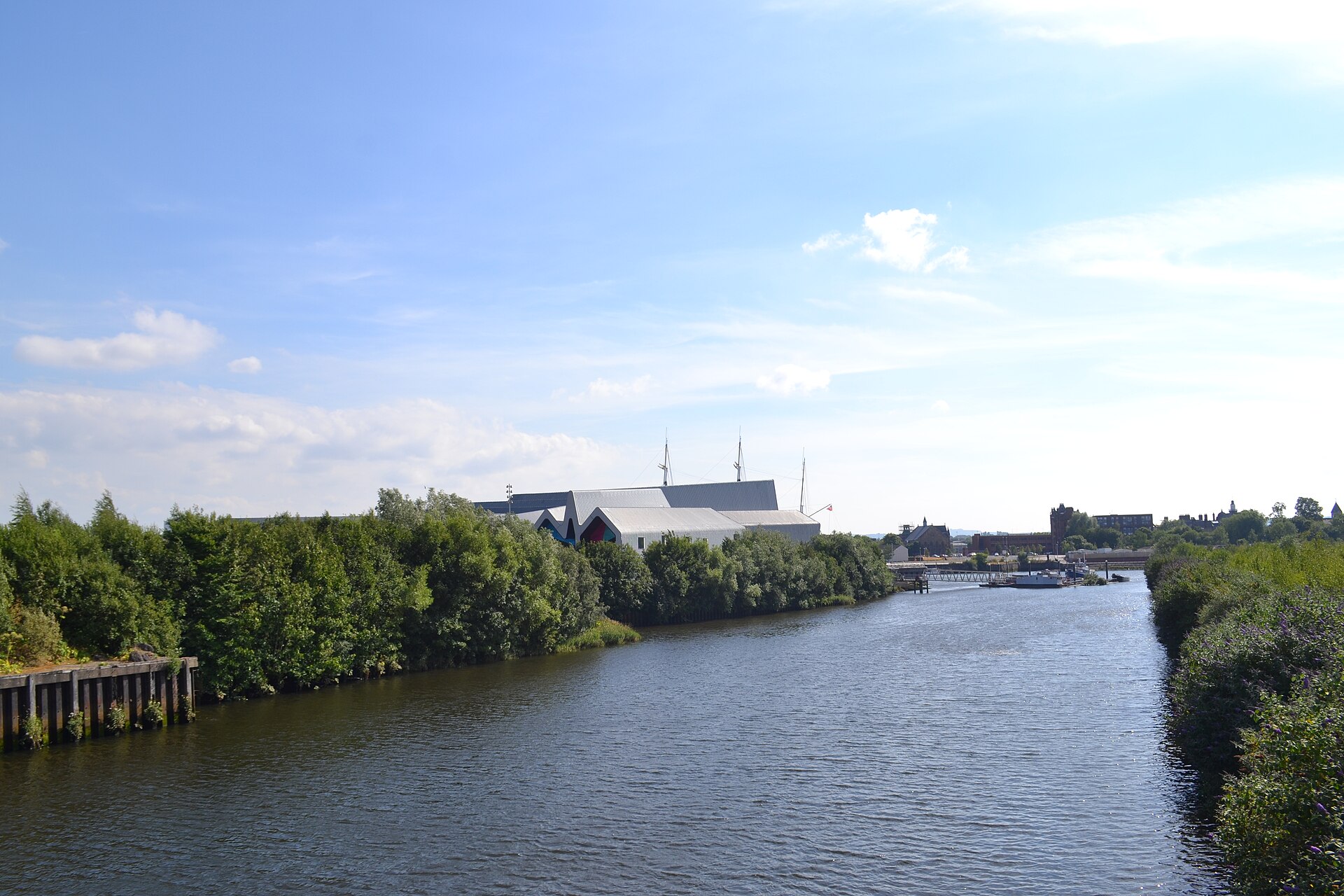 A river under a bright summer sky, with green banks and the zigzag roof of the Riverside Museum and the masts of the Glenlee beyond the trees
