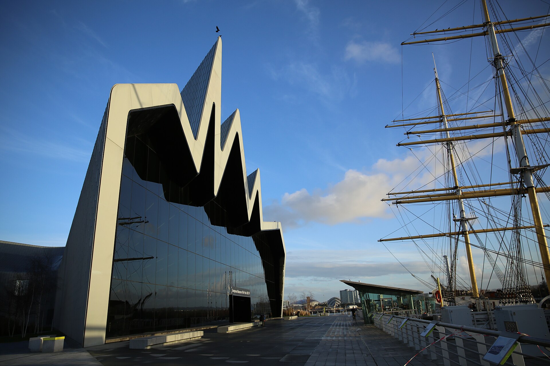 The zigzag roofline and glass front of the Riverside Museum on the Clyde, with the masts and rigging of the tall ship Glenlee on the right under a blue sky