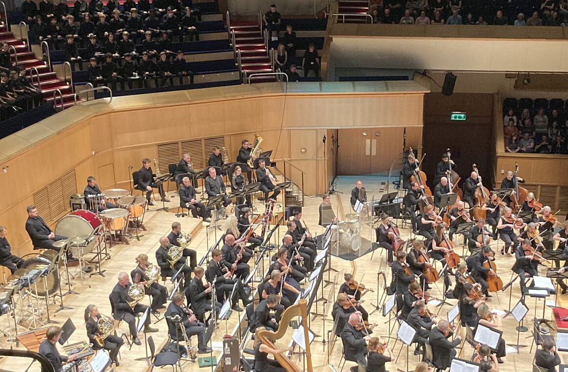 The Royal Scottish National Orchestra on a wide wooden stage seen from above, with the audience seated on tiers around the hall