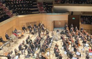 The Royal Scottish National Orchestra on a wide wooden stage seen from above, with the audience seated on tiers around the hall