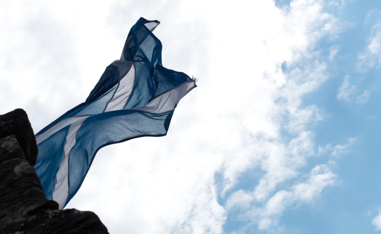 A blue and white Saltire flag streaming out in the wind against a bright, cloudy sky beside dark stonework