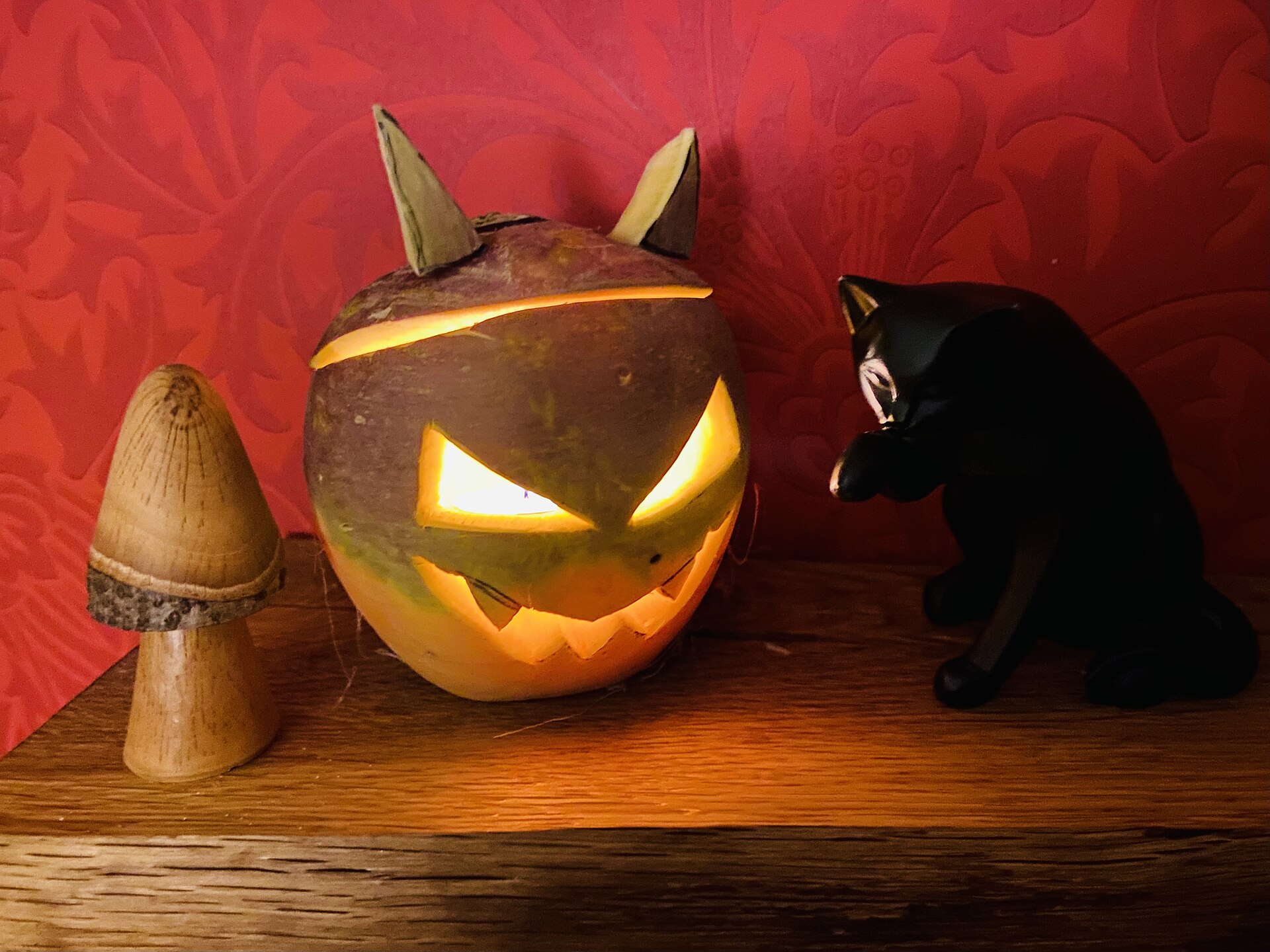 A carved turnip lantern with candlelight glowing through its slanted eyes and jagged grin, beside a wooden toadstool and a black cat ornament
