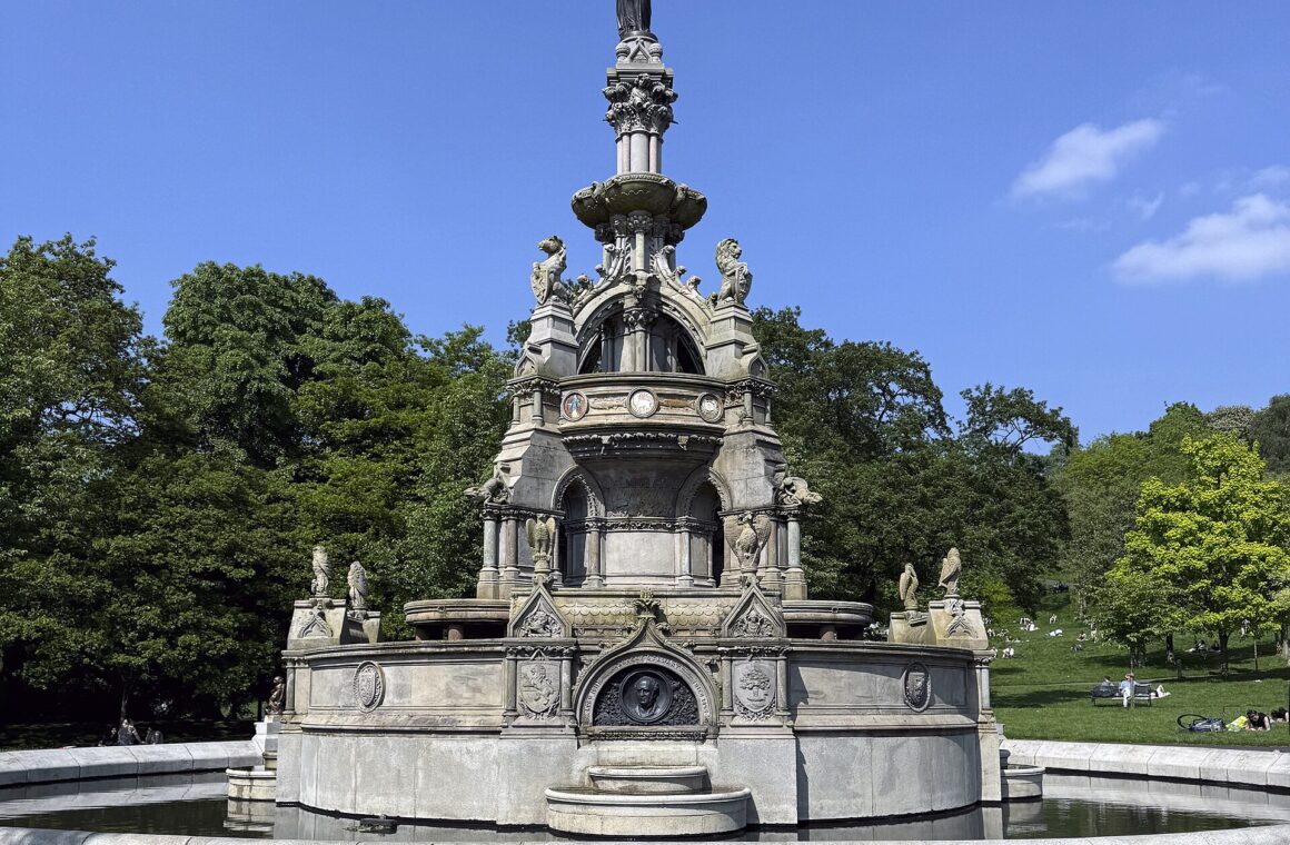 The ornate Stewart Memorial Fountain in Kelvingrove Park under a deep blue sky, with trees in fresh leaf and people relaxing on the grass