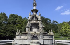 The ornate Stewart Memorial Fountain in Kelvingrove Park under a deep blue sky, with trees in fresh leaf and people relaxing on the grass