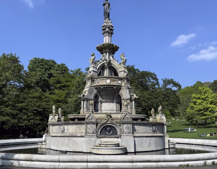 The ornate Stewart Memorial Fountain in Kelvingrove Park under a deep blue sky, with trees in fresh leaf and people relaxing on the grass