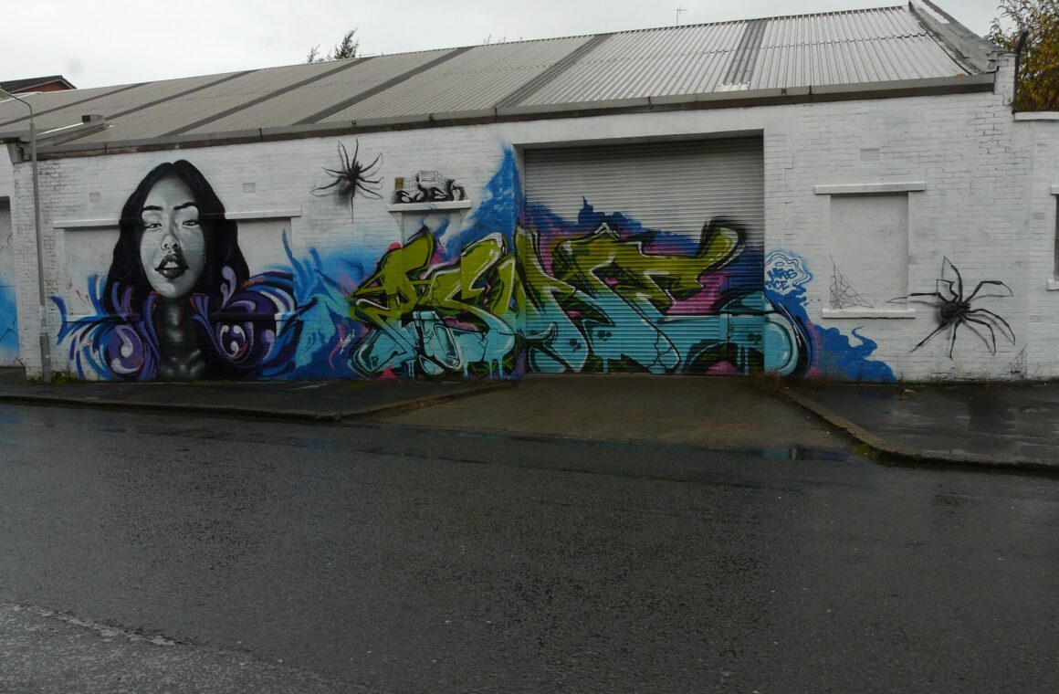 A long white warehouse wall on Eastvale Place painted with graffiti, including a portrait of a woman in blue and a large green tag