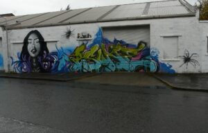 A long white warehouse wall on Eastvale Place painted with graffiti, including a portrait of a woman in blue and a large green tag