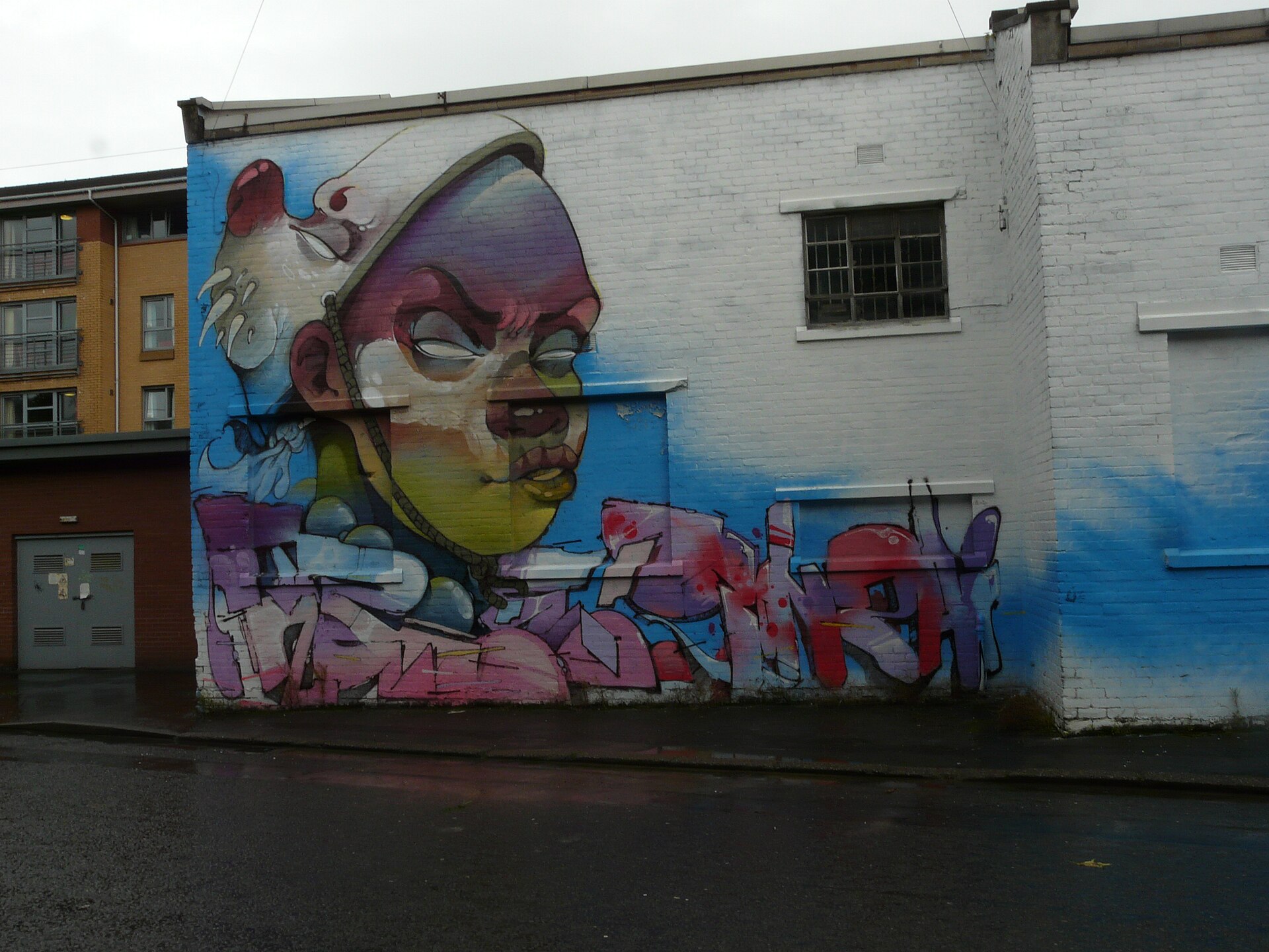A large graffiti mural of a stylised clown-like face in pink, blue and yellow painted across the white brick wall of a warehouse on Eastvale Place