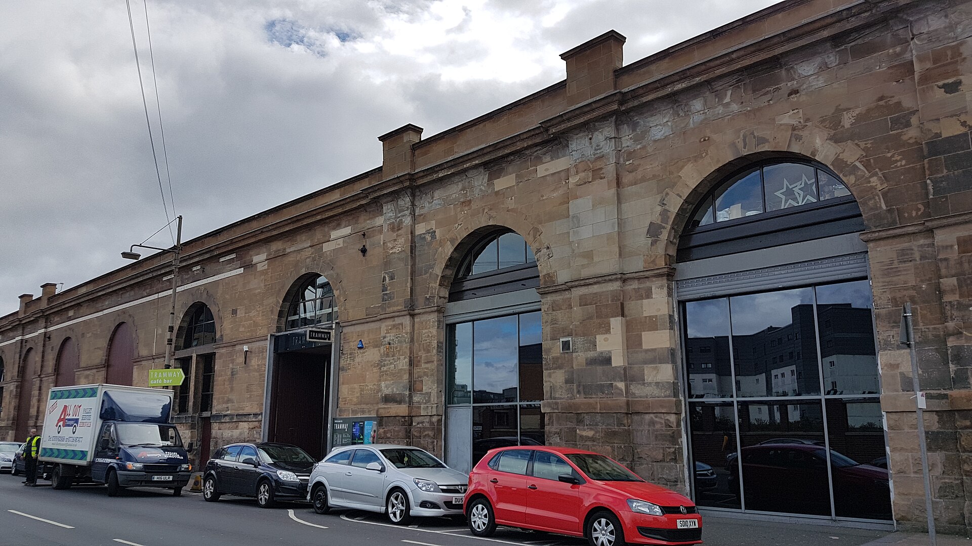 The long red sandstone frontage of Tramway on Albert Drive, with tall arched windows, a modern glass entrance and cars parked along the street