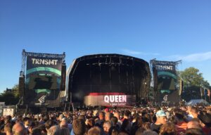 The TRNSMT main stage on Glasgow Green under a clear blue sky, with a large crowd in front of the stage and the headline act's name on the screen