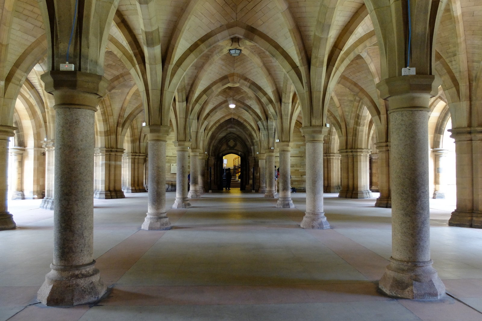 The vaulted Gothic cloisters beneath the University of Glasgow's main building, with rows of stone pillars and ribbed arches leading to a lit doorway