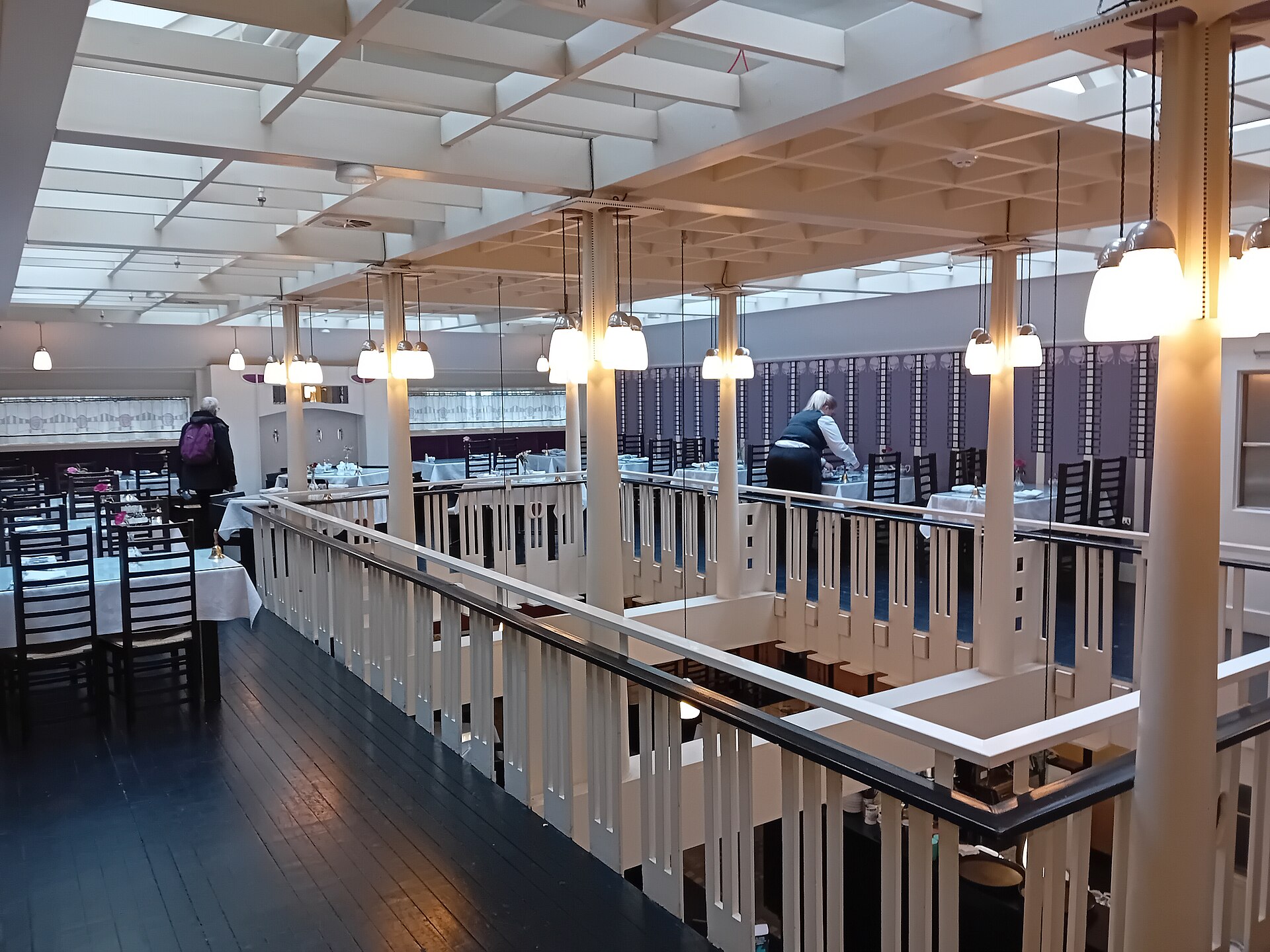 The upstairs gallery of the Willow Tearooms, with white Mackintosh balustrades and pillars, pendant lamps and tables laid with white cloths and high-backed black chairs