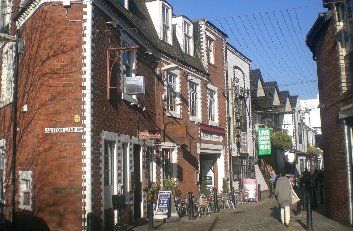 Ashton Lane in Glasgow's West End on a sunny day, with red-brick buildings, bar signs, outdoor tables and strings of lights overhead