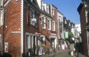 Ashton Lane in Glasgow's West End on a sunny day, with red-brick buildings, bar signs, outdoor tables and strings of lights overhead