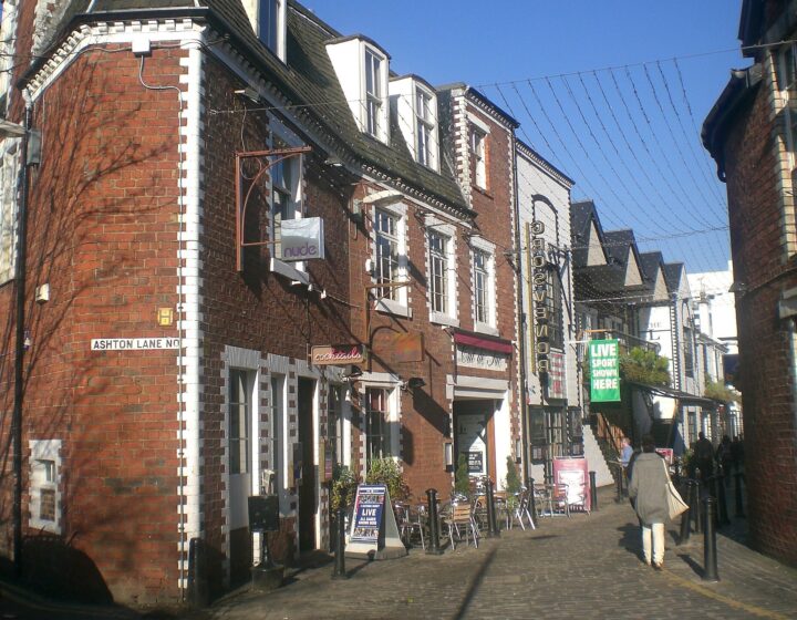 Ashton Lane in Glasgow's West End on a sunny day, with red-brick buildings, bar signs, outdoor tables and strings of lights overhead