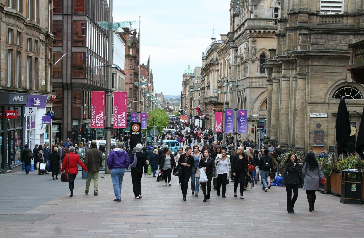 Crowds of shoppers walking along pedestrianised Buchanan Street between sandstone and modern buildings, with banners on lamp posts and the street rising into the distance