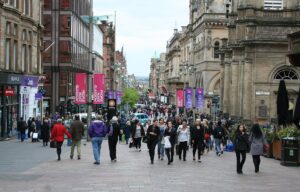 Crowds of shoppers walking along pedestrianised Buchanan Street between sandstone and modern buildings, with banners on lamp posts and the street rising into the distance