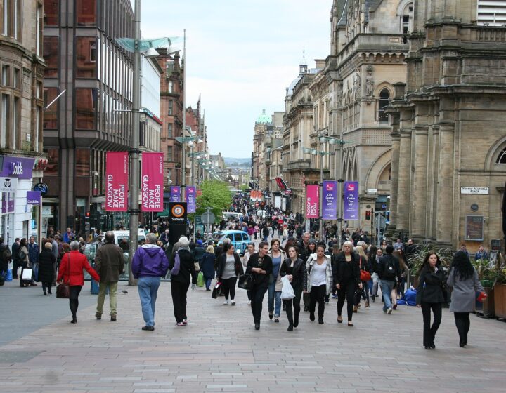 Crowds of shoppers walking along pedestrianised Buchanan Street between sandstone and modern buildings, with banners on lamp posts and the street rising into the distance
