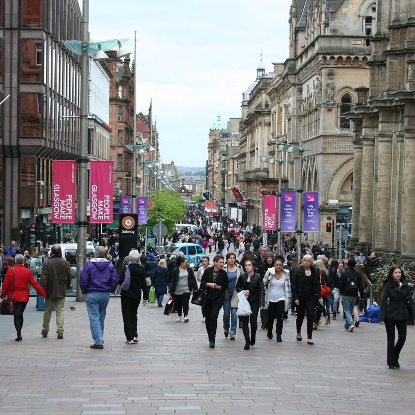 Crowds of shoppers walking along pedestrianised Buchanan Street between sandstone and modern buildings, with banners on lamp posts and the street rising into the distance
