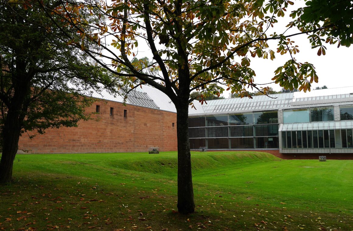 The red sandstone and glass Burrell Collection building beyond a sloping lawn and a horse chestnut tree in early autumn, in Pollok Country Park