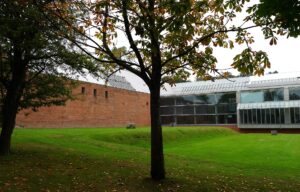 The red sandstone and glass Burrell Collection building beyond a sloping lawn and a horse chestnut tree in early autumn, in Pollok Country Park