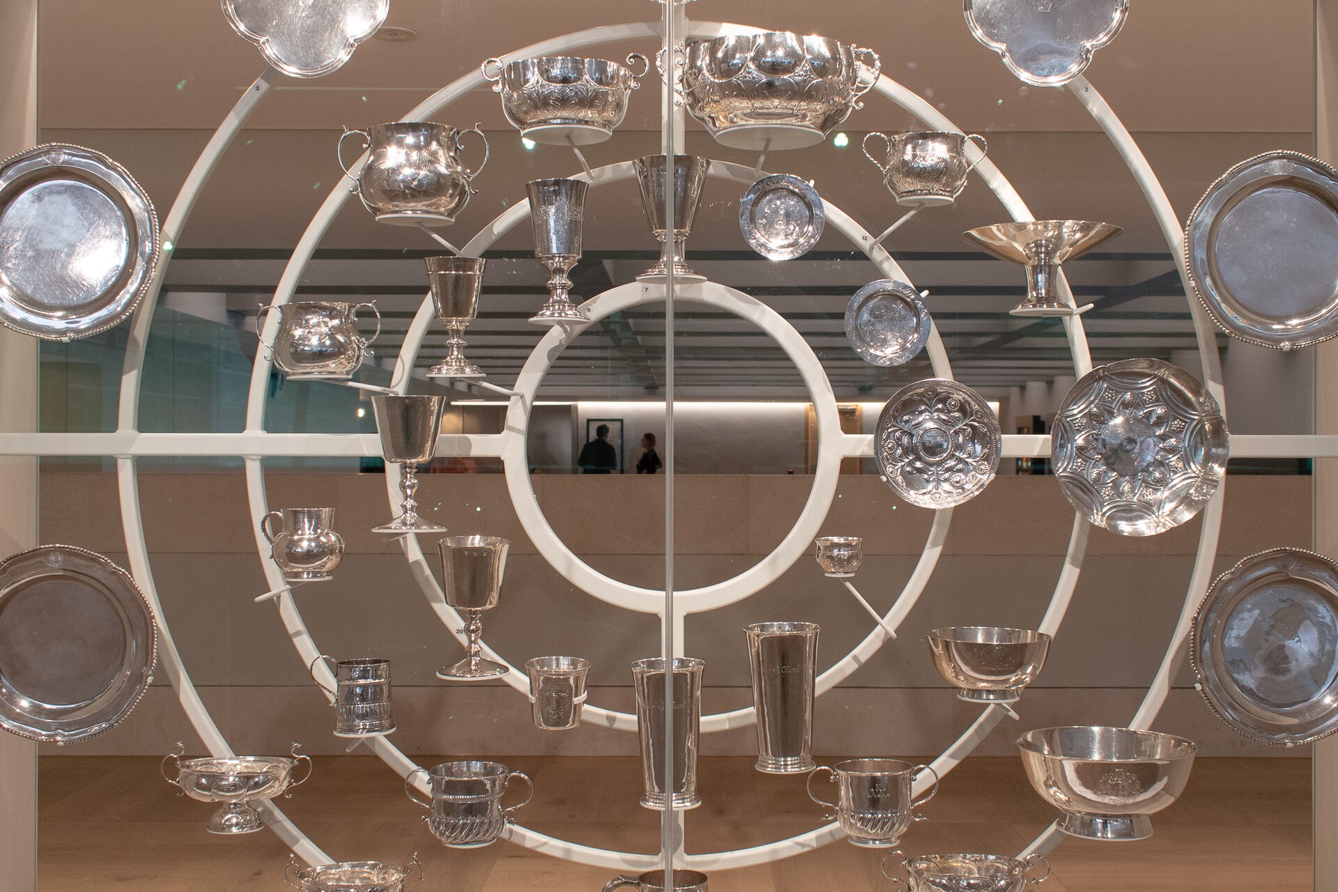 A large circular display of polished silver cups, tankards, bowls and plates mounted on white rings inside the refurbished Burrell Collection, with a gallery visible through the glass behind