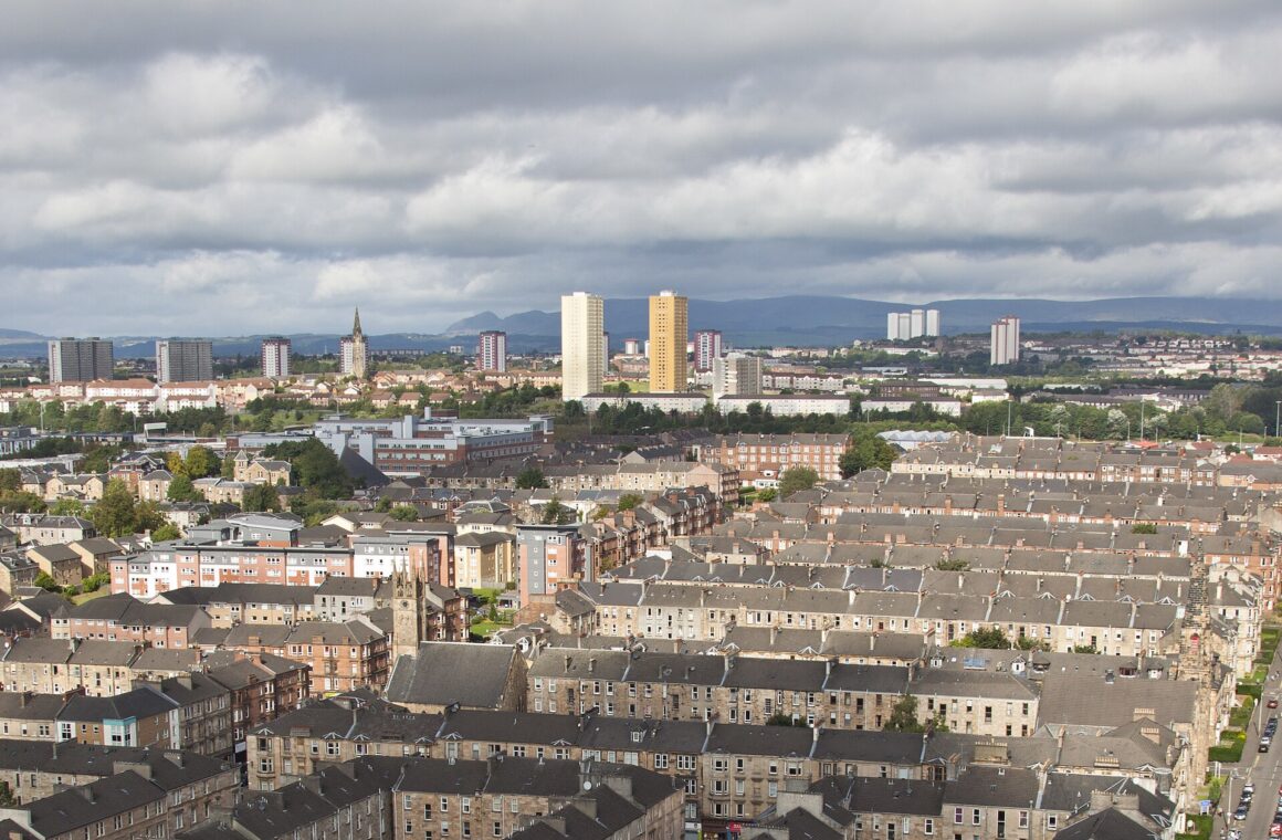Looking down over Dennistoun's grid of sandstone tenement blocks and back courts towards tower blocks and the Campsie Fells on the horizon under a cloudy sky