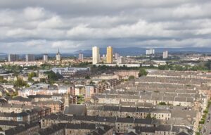 Looking down over Dennistoun's grid of sandstone tenement blocks and back courts towards tower blocks and the Campsie Fells on the horizon under a cloudy sky
