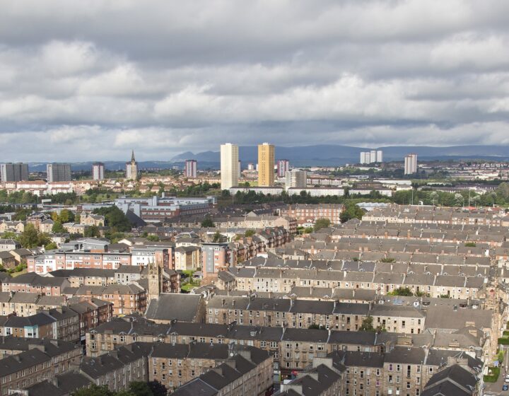 Looking down over Dennistoun's grid of sandstone tenement blocks and back courts towards tower blocks and the Campsie Fells on the horizon under a cloudy sky