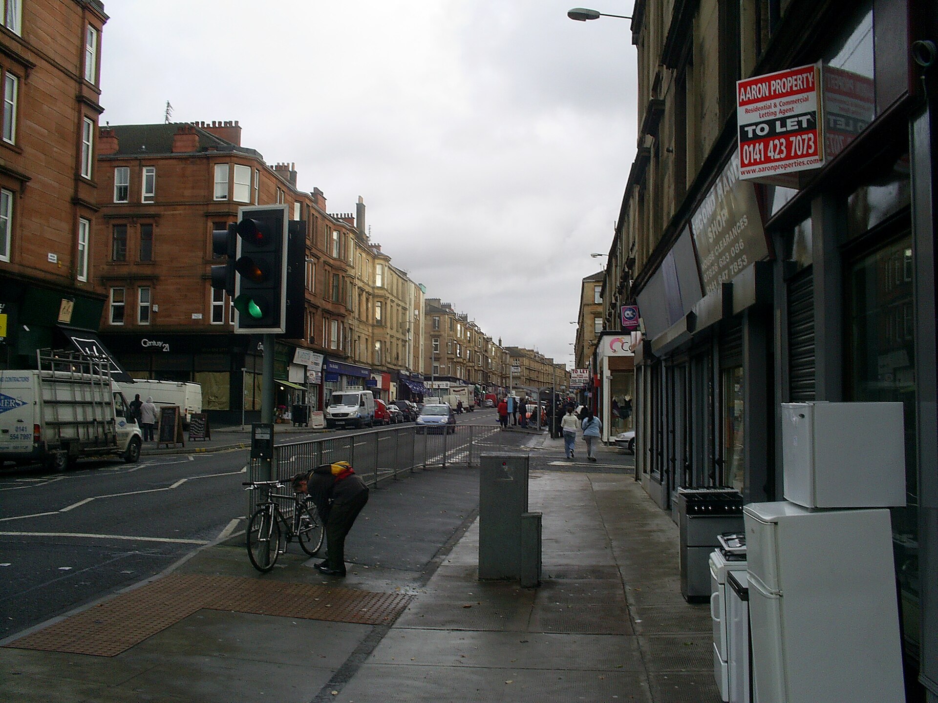 A long straight street lined with sandstone tenements and ground-floor shops, with traffic lights, a few pedestrians and parked vans under an overcast sky
