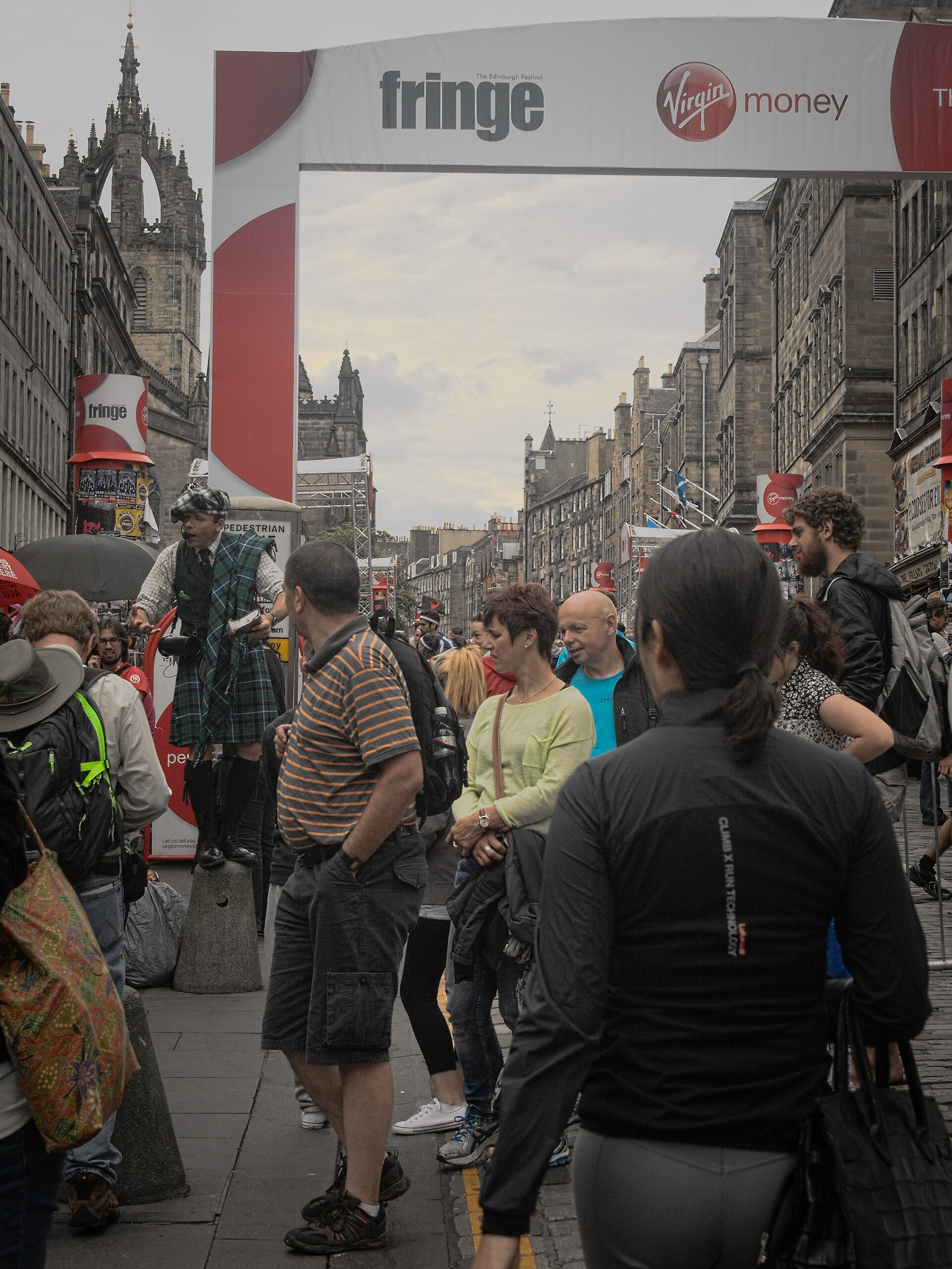 A street performer in a kilt raised above the crowd under a red and white Fringe banner stretched across Edinburgh's Royal Mile, with the crown spire of St Giles' Cathedral behind