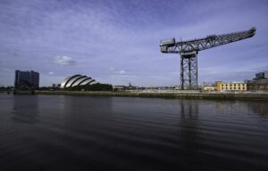 The Finnieston Crane standing over the River Clyde under a blue sky, with the silver shells of the Armadillo and a tall hotel block further along the quay, reflected in calm water