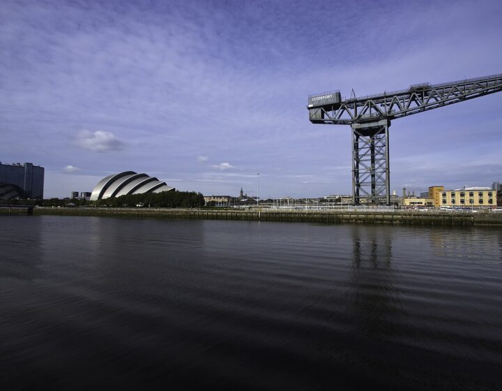 The Finnieston Crane standing over the River Clyde under a blue sky, with the silver shells of the Armadillo and a tall hotel block further along the quay, reflected in calm water