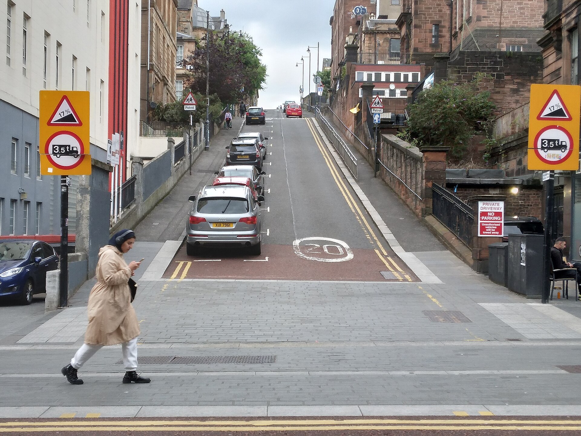 A steep street climbing away from a junction, with gradient warning signs on both sides, parked cars on the slope and a woman walking across the foot of the hill