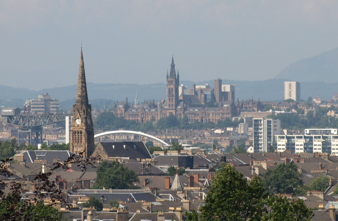 Looking north from Queen's Park over Southside tenement rooftops and a church spire towards the University of Glasgow tower, the Clyde Arc, the Finnieston Crane and hills on the horizon