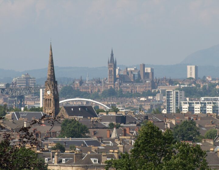 Looking north from Queen's Park over Southside tenement rooftops and a church spire towards the University of Glasgow tower, the Clyde Arc, the Finnieston Crane and hills on the horizon