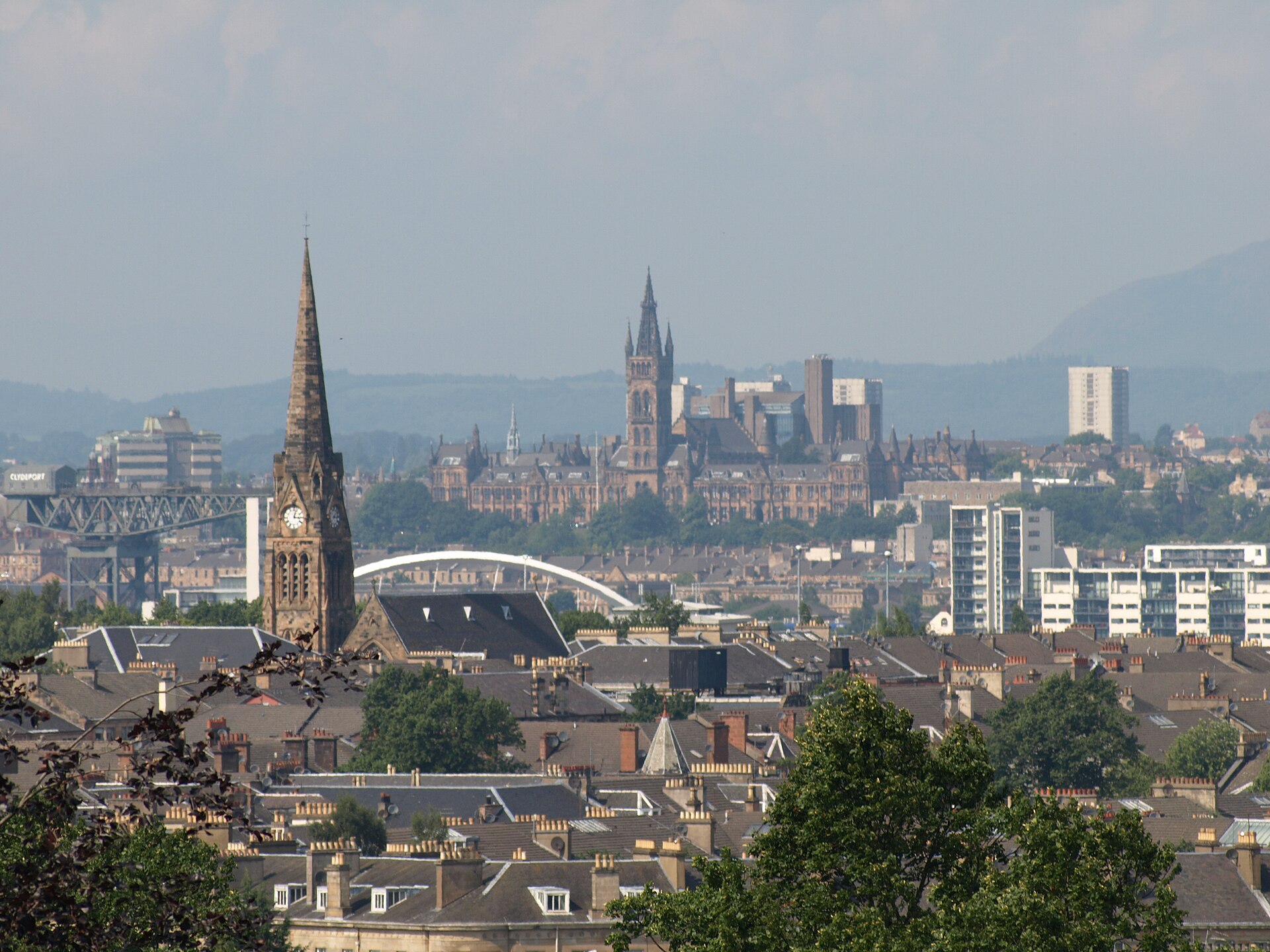 Looking north from Queen's Park over Southside tenement rooftops and a church spire towards the University of Glasgow tower, the Clyde Arc, the Finnieston Crane and hills on the horizon