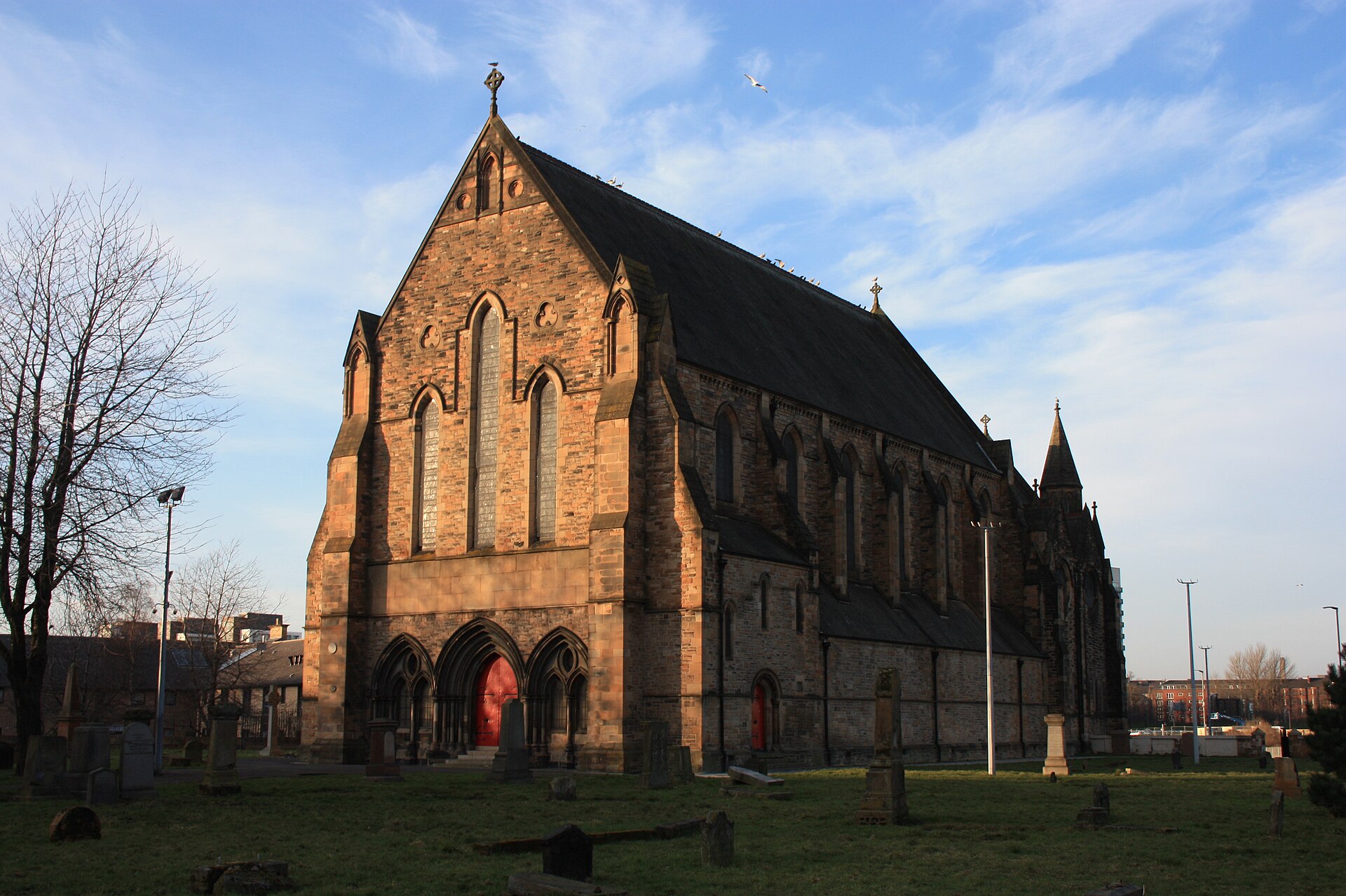 The red sandstone Govan Old Parish Church in low evening sun, with its Gothic windows and red doors, set in a grassy churchyard of old gravestones