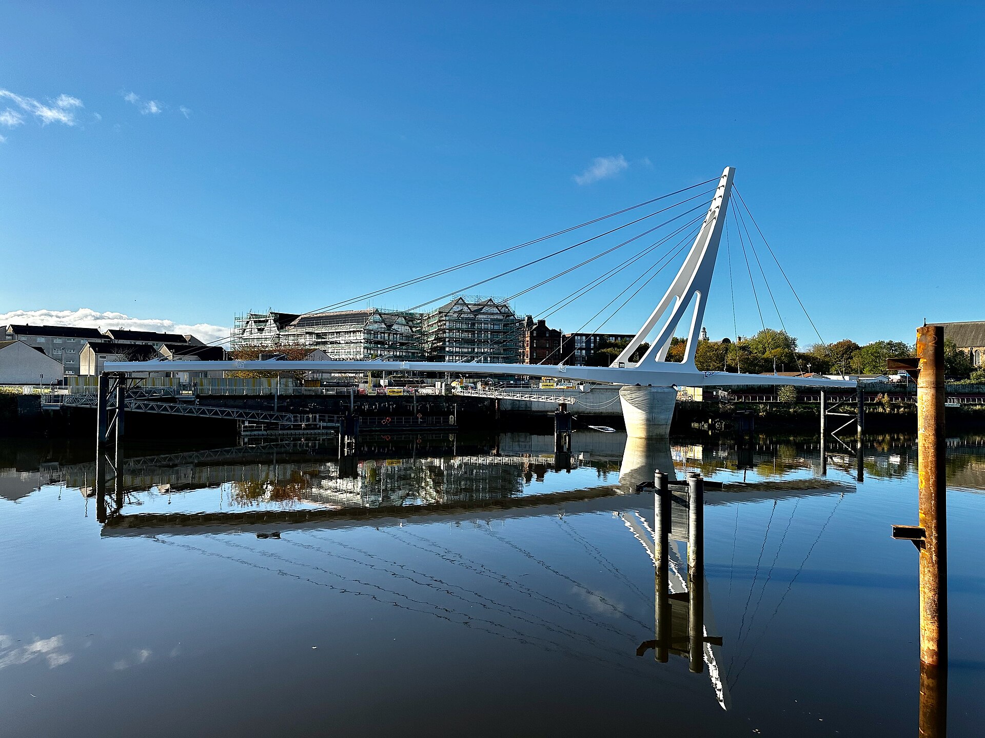 A white cable-stayed footbridge spanning a calm river under a clear blue sky, its mast leaning towards the far bank, where new flats stand beside older sandstone buildings