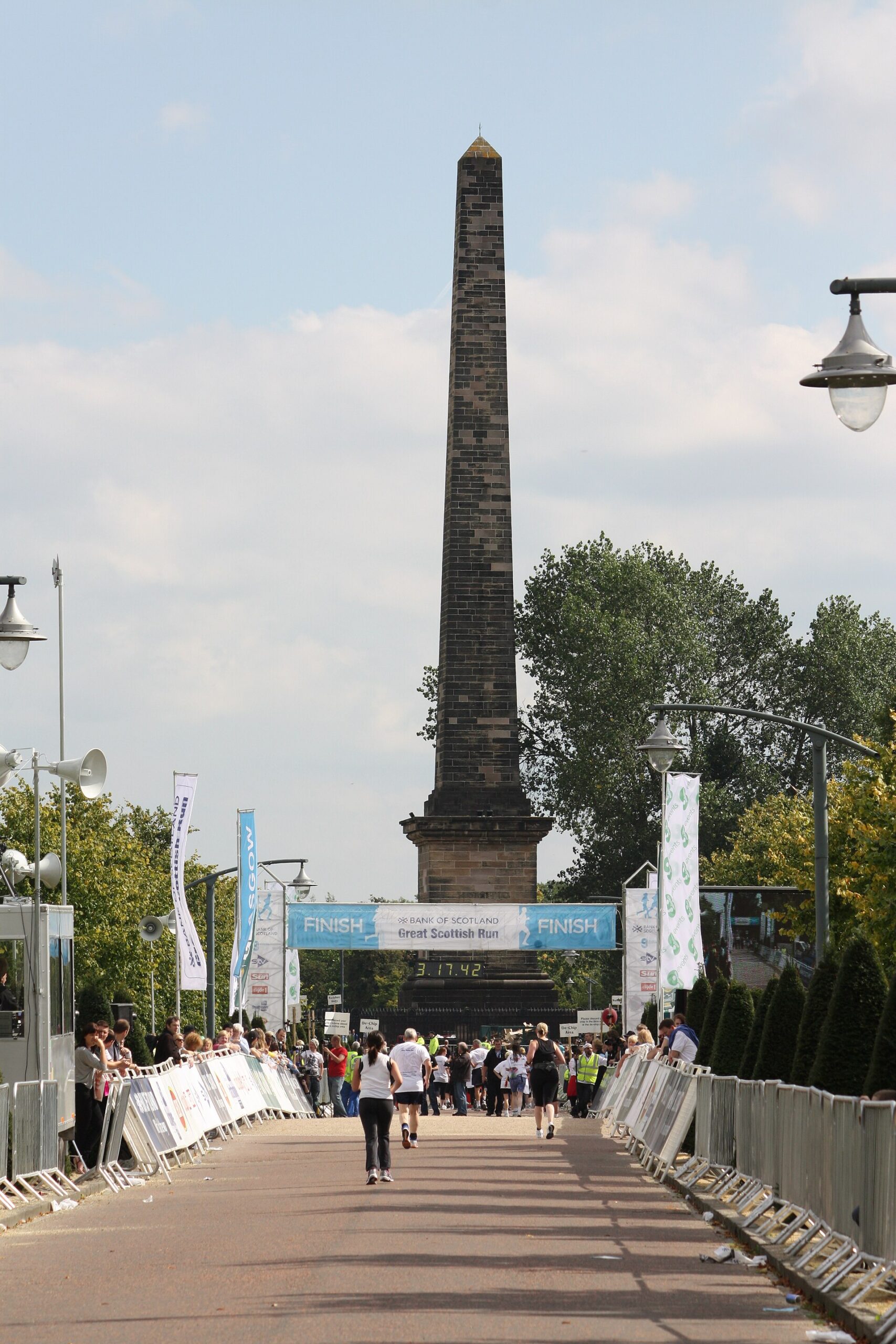 Runners heading down a barriered avenue towards a blue Finish arch on Glasgow Green, with the tall stone obelisk of the Nelson Monument behind