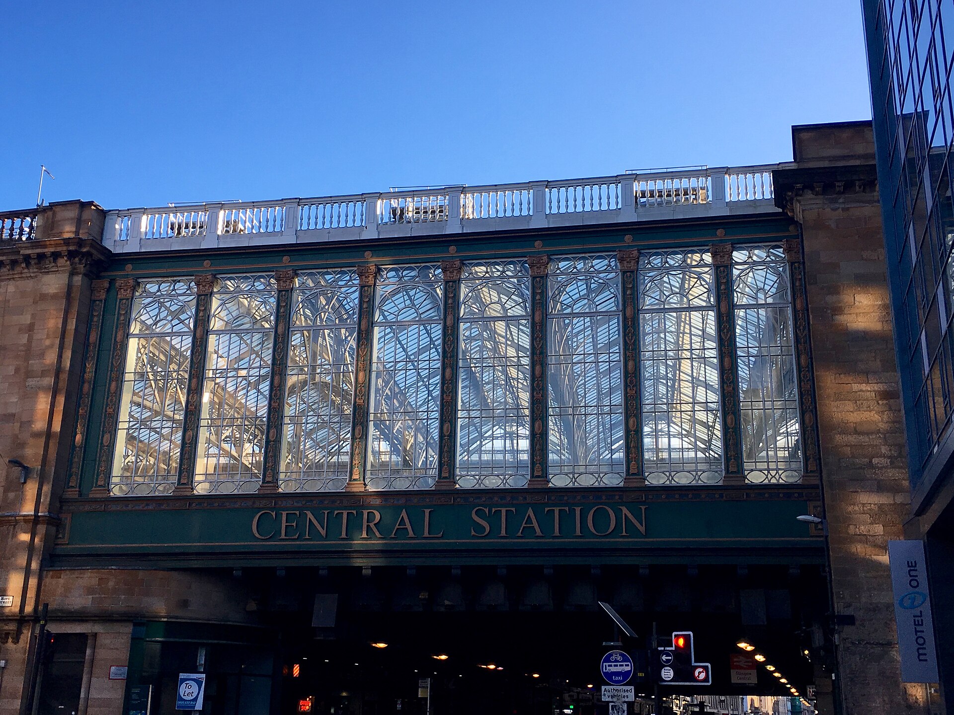 The glass-walled railway bridge of Glasgow Central Station spanning Argyle Street, with tall arched windows above a green iron fascia lettered Central Station, under a clear blue sky