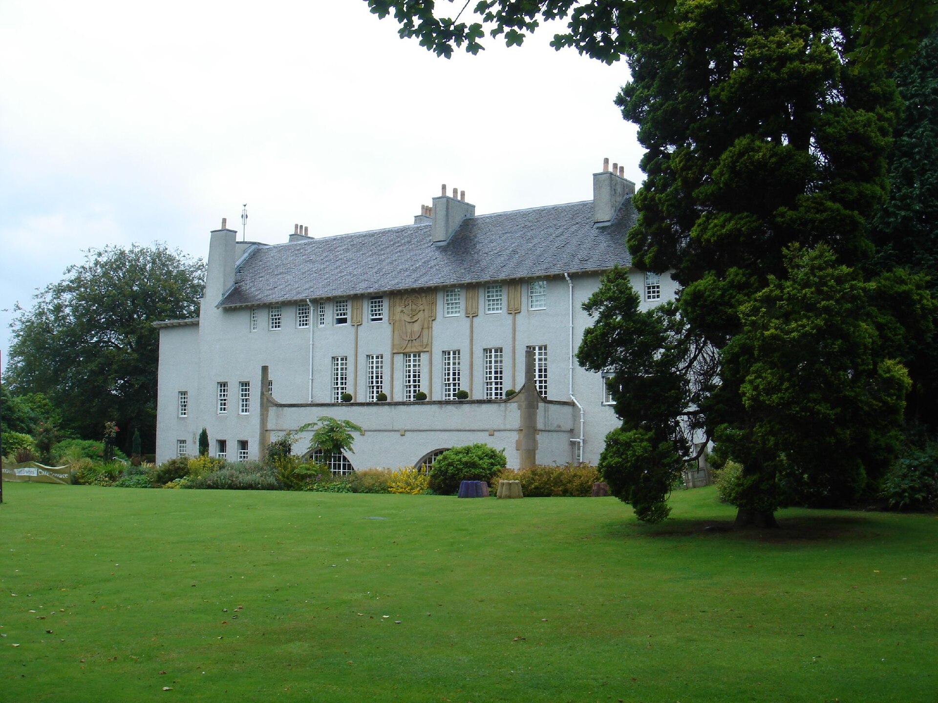 The white harled House for an Art Lover with tall windows, carved panels and a slate roof, seen across a wide lawn with trees and flower beds