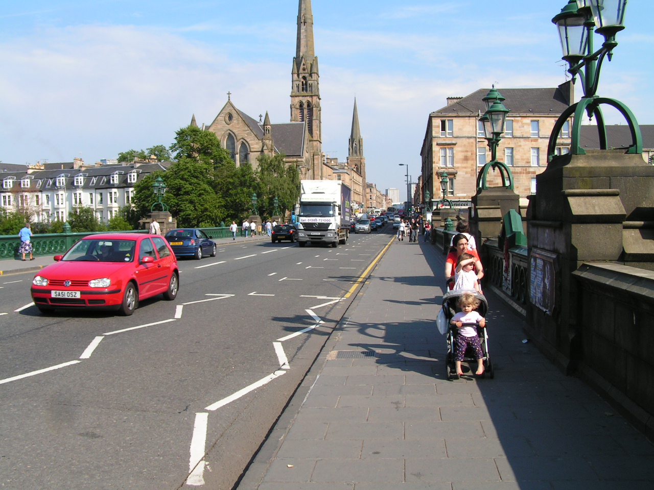 Great Western Road crossing the River Kelvin on an ornate green iron bridge with lamp standards, looking west towards a tall church spire and sandstone terraces on a sunny day