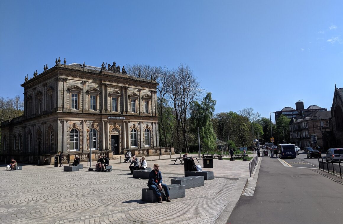 The Italianate sandstone Langside Hall behind the paved square at Shawlands Cross on a sunny day, with people sitting on stone benches and tenements beyond