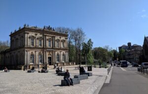 The Italianate sandstone Langside Hall behind the paved square at Shawlands Cross on a sunny day, with people sitting on stone benches and tenements beyond