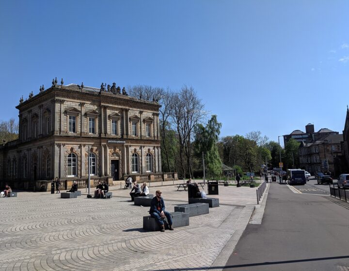 The Italianate sandstone Langside Hall behind the paved square at Shawlands Cross on a sunny day, with people sitting on stone benches and tenements beyond