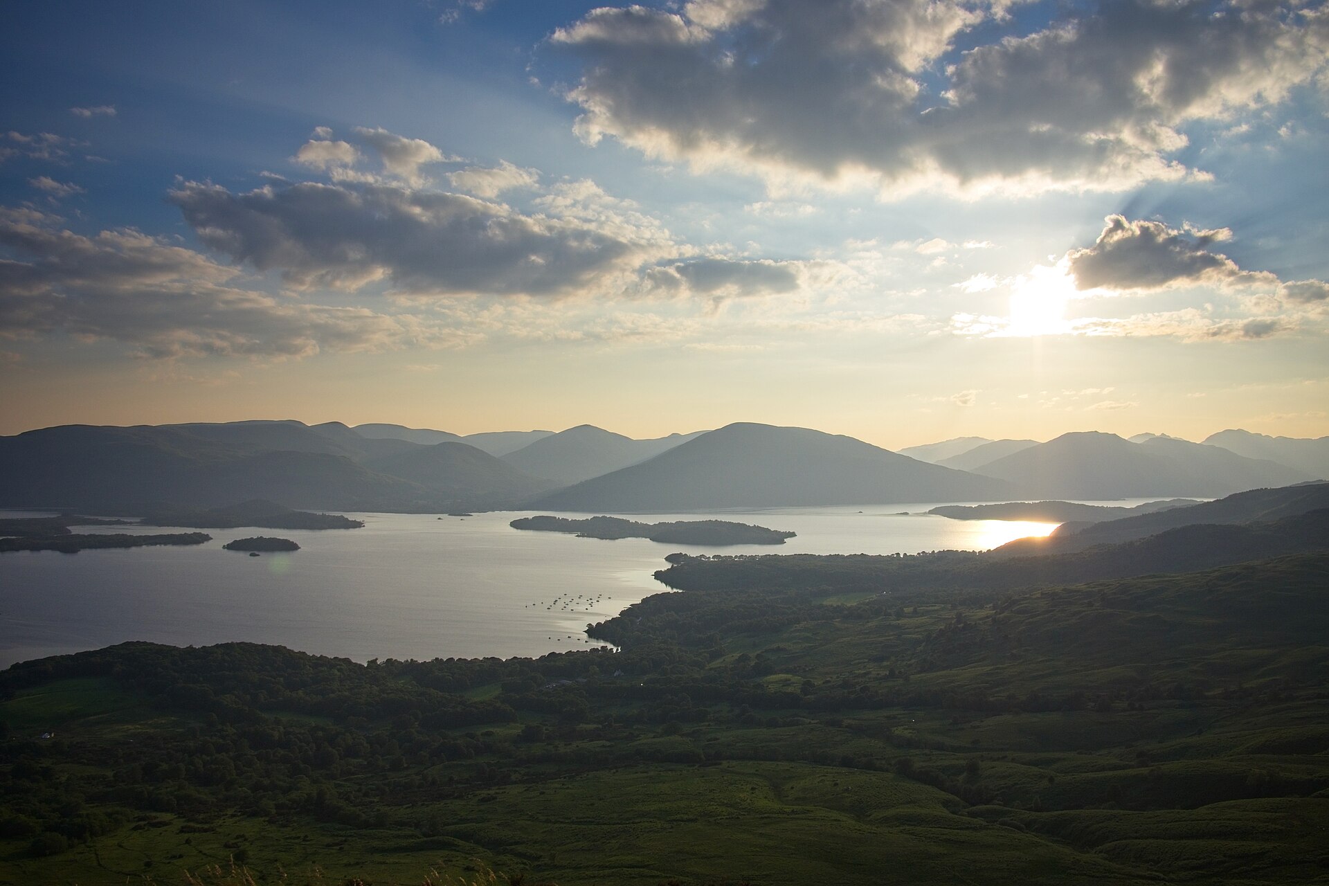 Loch Lomond seen from the slopes of Conic Hill in low sun, with a line of wooded islands crossing the loch and mountains beyond under broken cloud