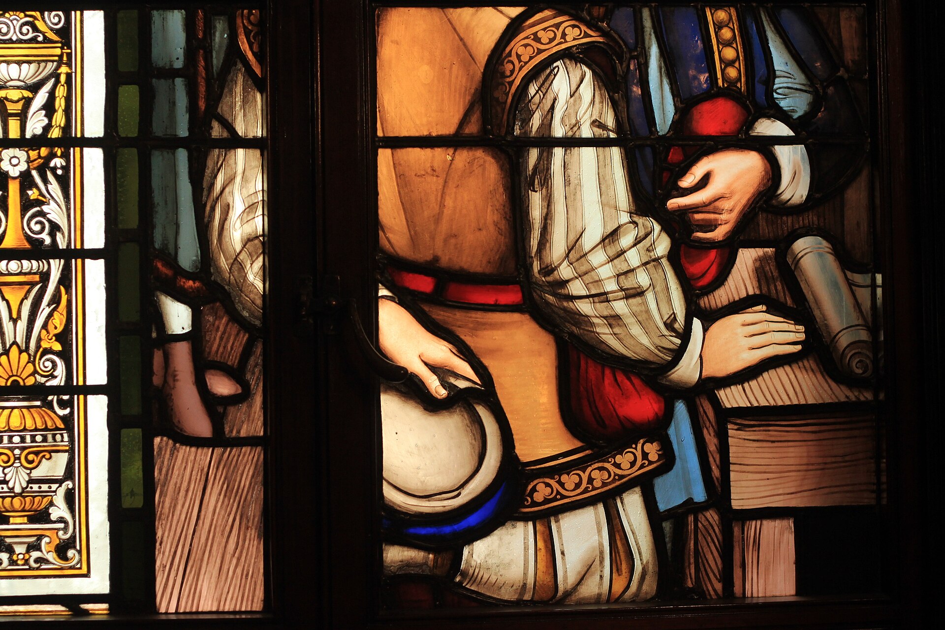 Close-up of a richly coloured stained-glass window showing a figure in a puffed-sleeve tunic holding a rolled document, framed by ornate painted borders