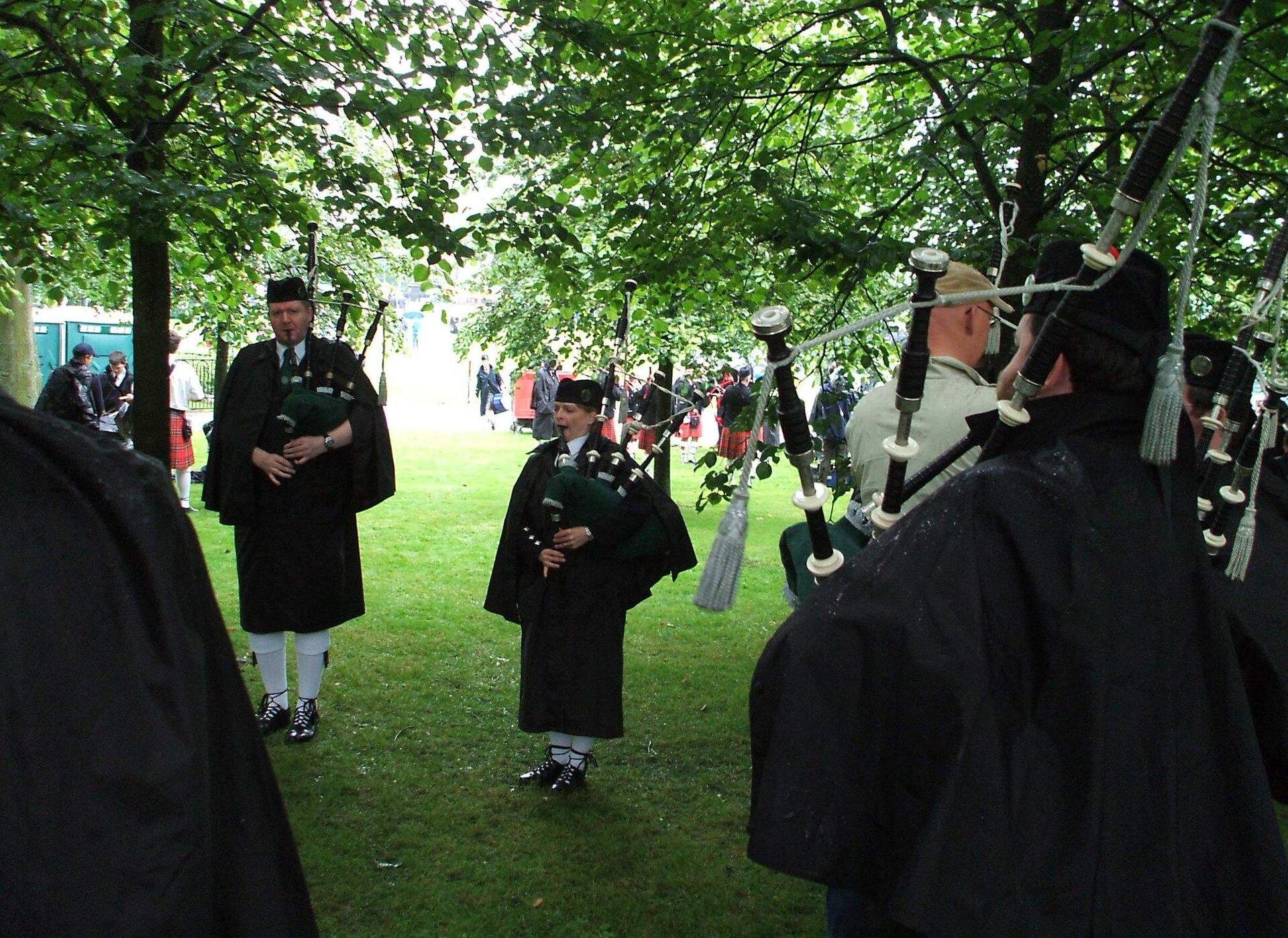 Pipers in long black rain capes tuning their bagpipes on wet grass under a canopy of trees, with other bands and spectators in the background